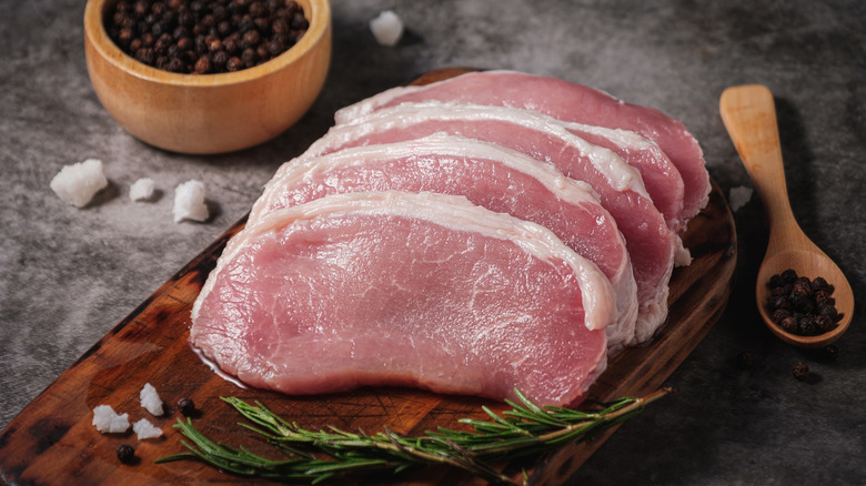 raw pork chops on a wooden board, with black peppercorns in a wooden bowl and spoon, surrounded by large salt rocks and a sprig of rosemary