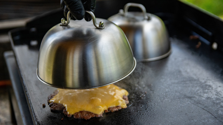 a black-gloved hand lifting a lid over a smashed burger on a griddle, covered in melted cheese