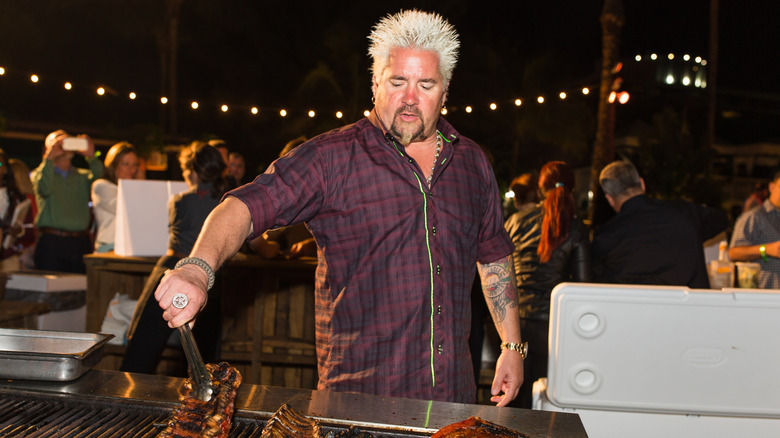 Guy Fieri wearing a purple check shirt, turning ribs on an outside grill