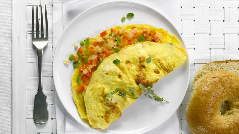 a white plate of scrambled eggs with veggies, between silver fork and bagel