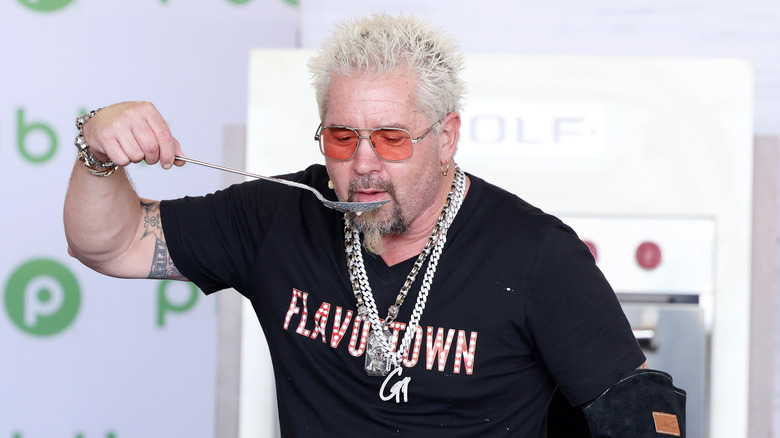 Guy Fieri wearing a black t-shirt with the word Flavortown on the front, eating from an oversized spoon