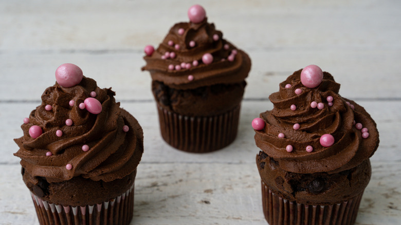 three chocolate cupcakes, decorated with chocolate buttercream and pink balls, on a white surface