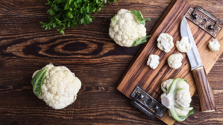 two small cauliflowers on a wooden surface, next to a small chopping board with florets on and knife