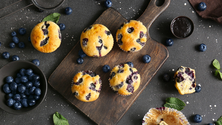 Blueberry muffins on a small wooden chopping board, surrounded by blueberries