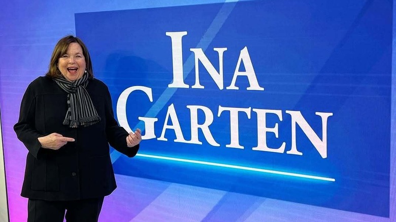 Ina Garten standing in a TV studio pointing and smiling at a graphic of her name in white writing on a blue background