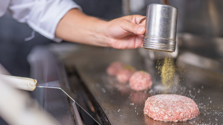 A person shaking seasoning on burgers over the grill