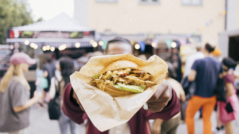 man with face obscured holding out a delicious sandwich to the camera, food stalls and people in background