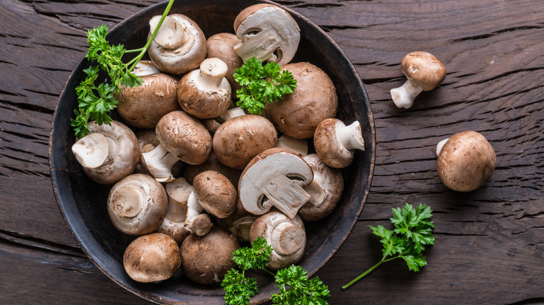 Top down view of cremini mushrooms in a dark bowl on wooden table with parsley