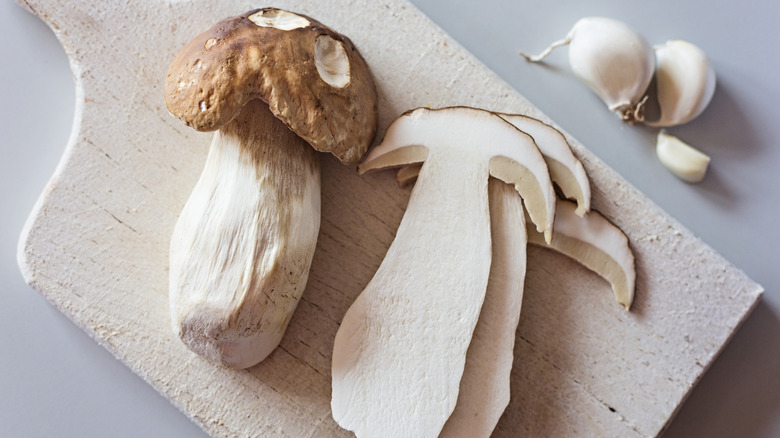 Whole and sliced porcini mushrooms on a white chopping board, next to three cloves of garlic