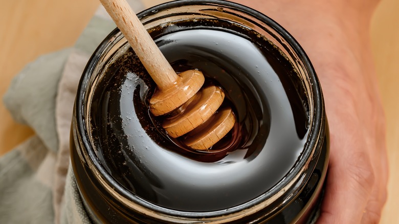 A hand holding a glass jar of buckwheat honey with a honey dipper pushed into it