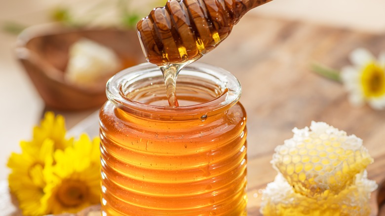 a wooden honey stick dripping golden honey into a glass jar, sitting next to a honey comb