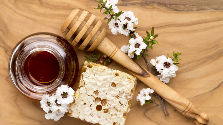 Looking down on a glass jar of Mānuka honey, next to a honey dipper, a honeycomb square and Mānuka flowers