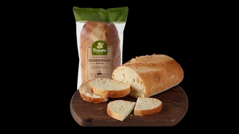 A bag of Panera Bread's sourdough bread, behind a loaf with three slices cut from it, on a wooden chopping board on a black background