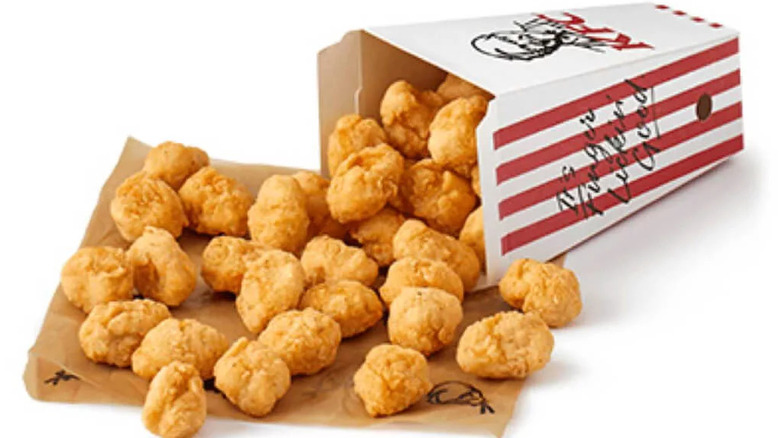 A box of popcorn chicken on its side, with several nuggets spilling onto a brown paper KFC bag