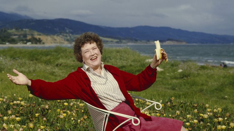 Julia Child sitting in nature eating a banana and smiling