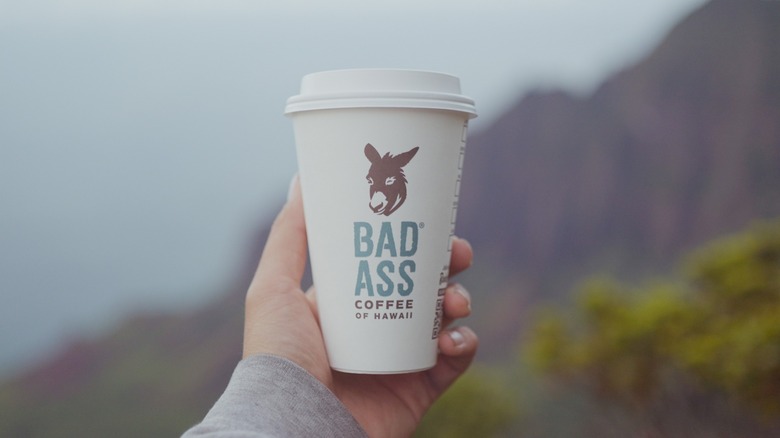 Hand holding branded cup of Bad Ass Coffee against blurred mountain background.