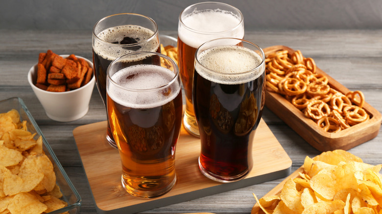 Overhead of different glasses of beer on wood board, surrounded by snacks.