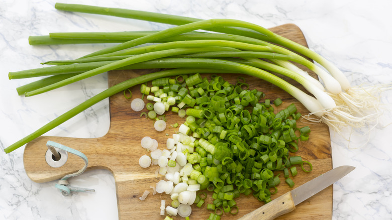 Whole and chopped green onions on wood cutting board with knife and marble counter.