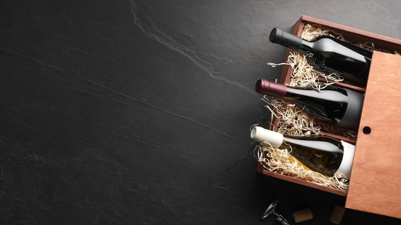 Three wine bottles in partially opened wooden case with straw, on dark slate background.