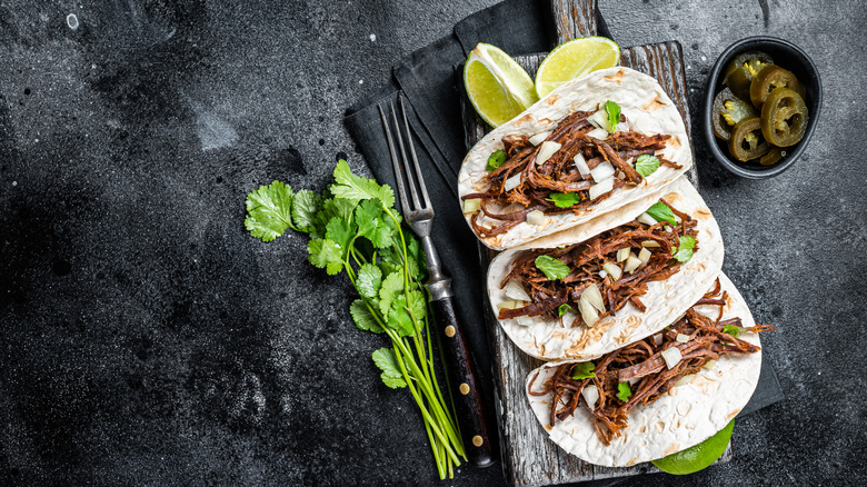 Three pork carnitas tacos on wood cutting board and black surface