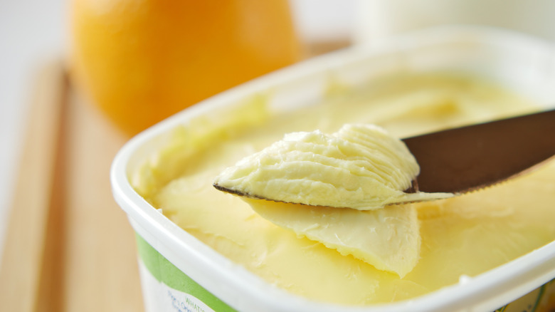 A close-up image of a tub of white tub of margarine with a green stripe. A butter knife laden with margarine is balanced on the side, and there is a soft, warm-toned blur on the background.