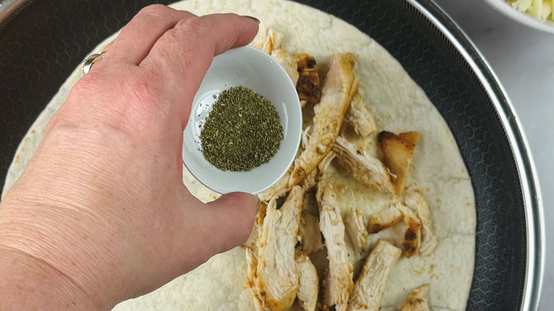 Hand holding bowl of dried herbs over chicken in skillet