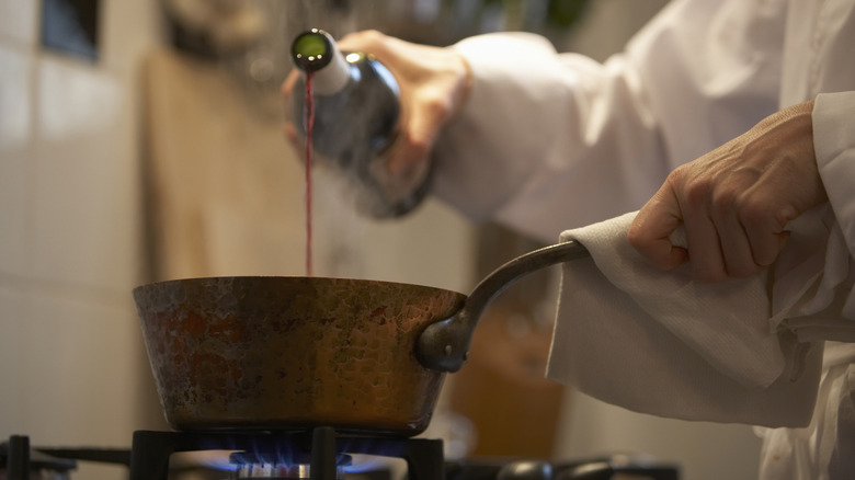 Person pouring red wine from bottle into copper pot on stove.