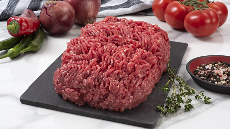 Raw ground beef sits on a black cutting board on a white surface alongside peppers, red onions, tomatoes, a sprig of thyme, and a bowl of whole peppercorns.