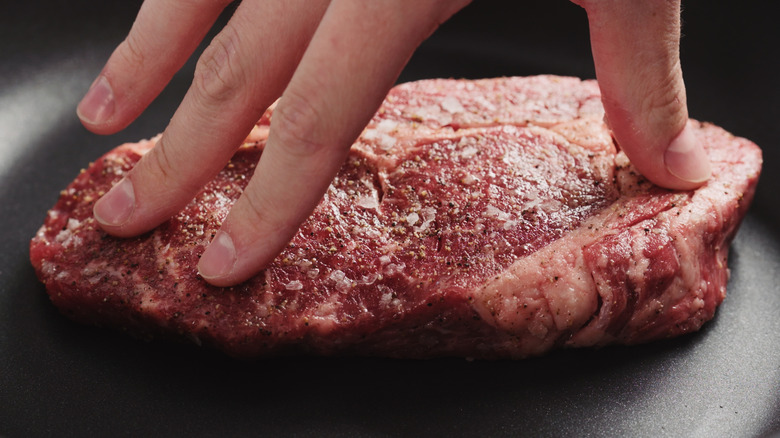 closeup of fingers pressing ribeye steak on non stick pan