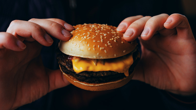 Hands holding cheeseburger against dark background.