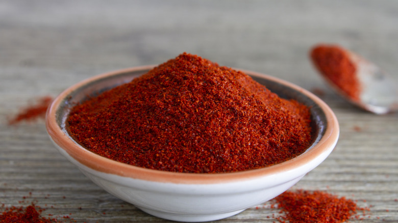 Sweet red Hungarian paprika is heaped high in a glazed ceramic bowl with an orange lip. A spoon sits in the blurred background