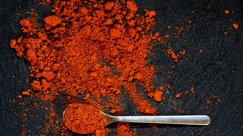 Vibrant, red-orange paprika is scattered on a black tabletop with a silver spoon