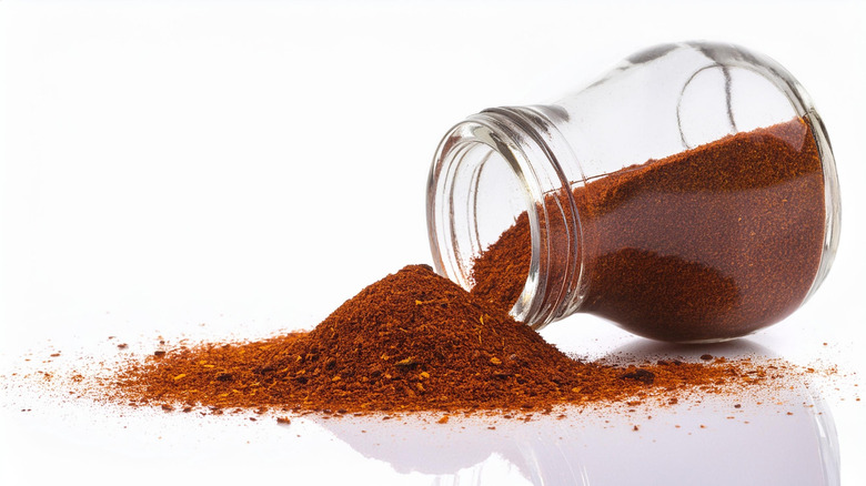 Brick red smoked paprika is spilled from a glass jar on a white surface, making a heap