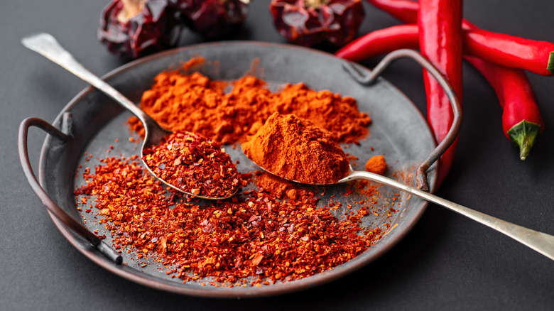 Spicy Spanish paprika sits on a metal plate alongside red chili flakes, with two silver spoons facing opposite directions and heaped in the spices. Fresh and dried red peppers are in the background on a black surface