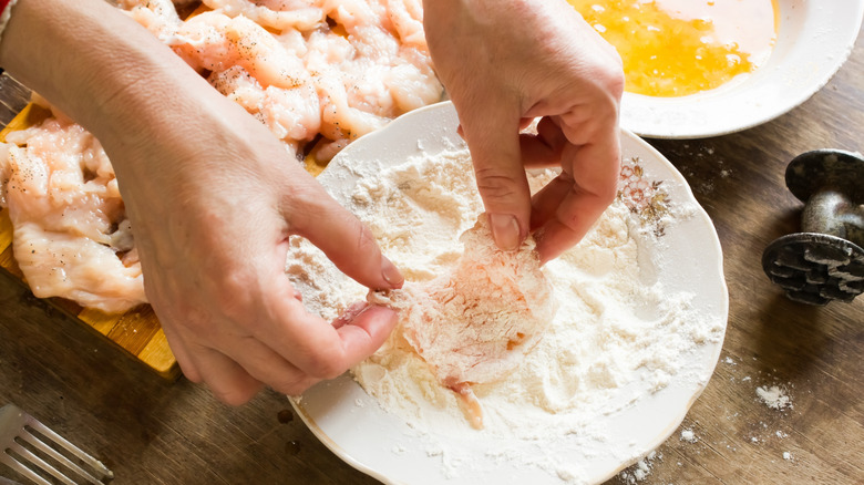 close up of hands dipping raw chicken meat in a plate of flour