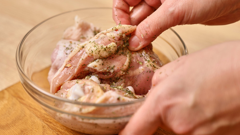 Hands lifting up a chicken breast in a glass bowl, marinating in spices and salt