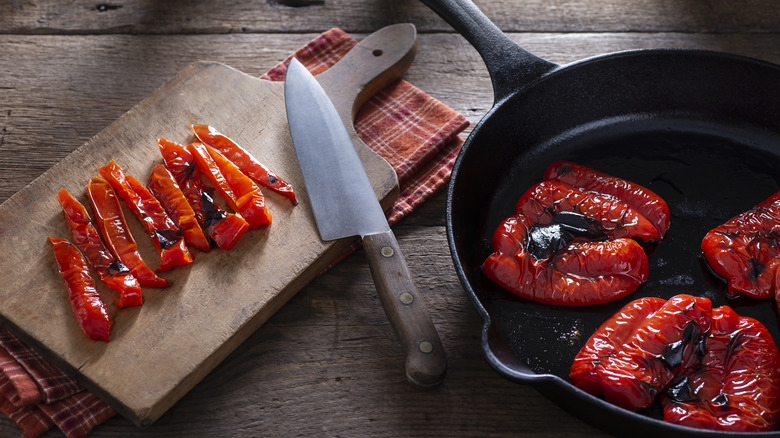 Sliced roasted red bell pepper on wood board next to whole roasted peppers in skillet.