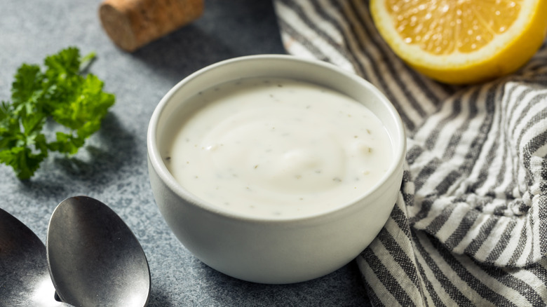 Ranch dressing in white ramekin along spoon, striped towel, parsley, and lemon half.