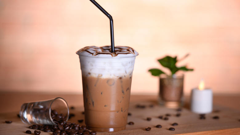 A frozen frappe on a table surrounded by coffee beans with a plant and candle in the background.