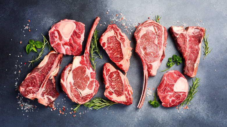 An assortment of fresh, juicy raw steaks are arranged end-to-end on a black surface, sprinkled with flaky salt and garnished with green herbs like rosemary, basil, and parsley.