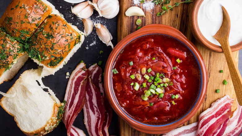 Borscht in ceramic bowl, surrounded by fresh rolls, cured meat, garlic, and salt