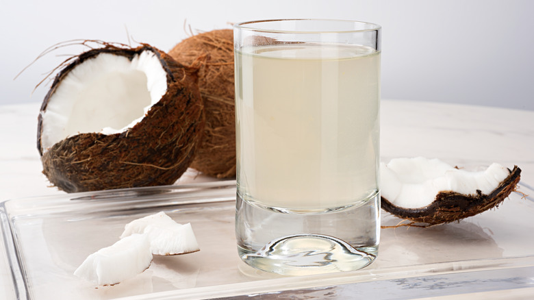 A glass of coconut water with cracked coconuts in the background.