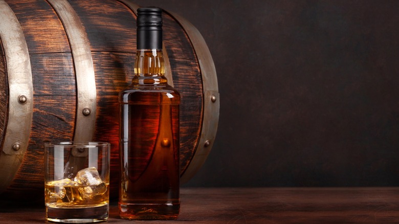 Bourbon bottle next to rocks glass with bourbon in front of wood barrel