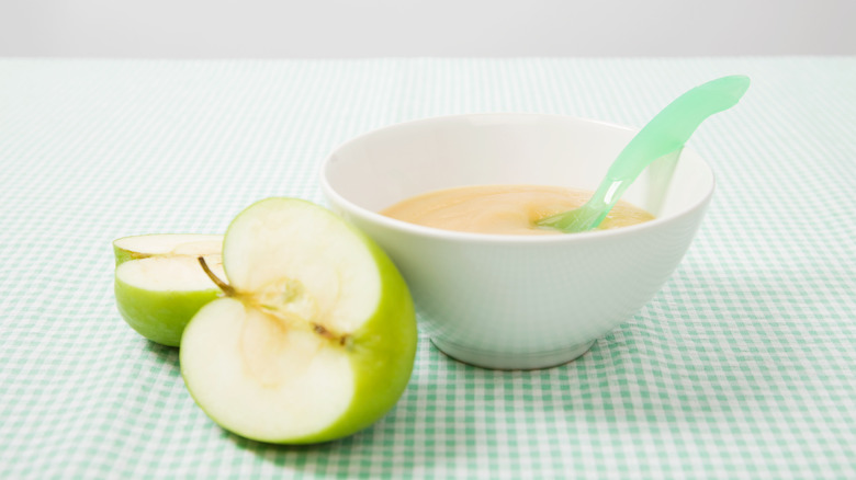 A white ceramic dish of applesauce with a green plastic spoon sticking out is set on a green gingham tablecloth alongside some cut green apples.