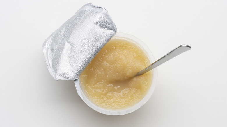 An individual serving of applesauce in a plastic cup with a peeled-back foil lid sits against a white background, a silver spoon sticking out.