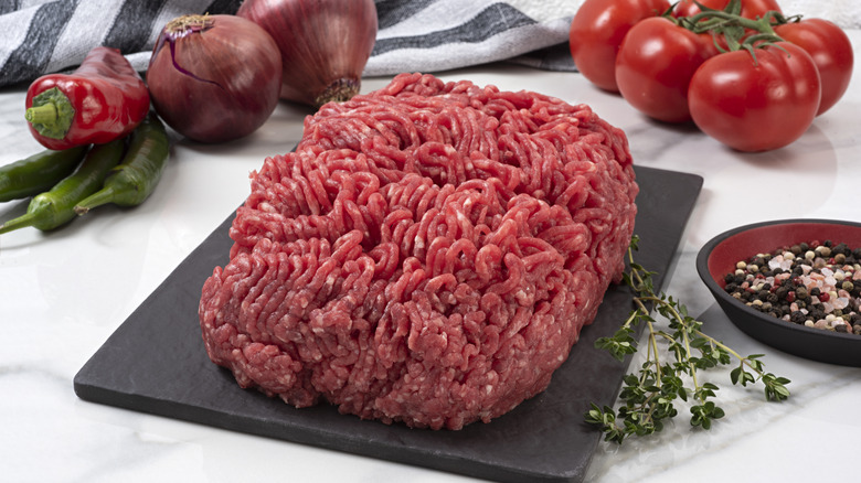 A pound of ground beef on a cutting board, surrounded by onions, peppers, seasonings, and tomatoes.