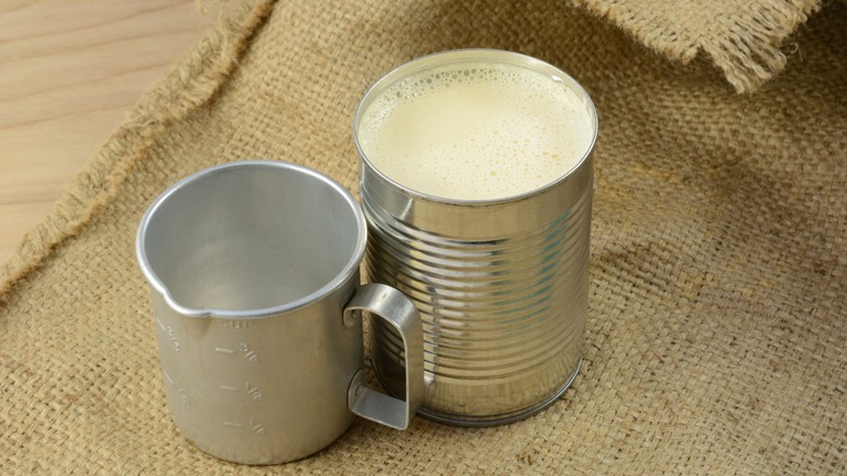 Opened can of evaporated milk next to vintage measuring cup on burlap