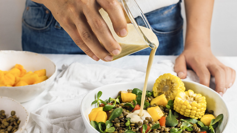 A woman pouring tahini over a salad greens, corn, lentils, and other vegetables.