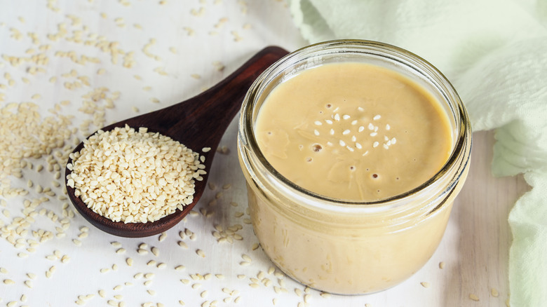 Top view of a jar of tahini topped with and surrounded by sesame seeds with wood spoon.