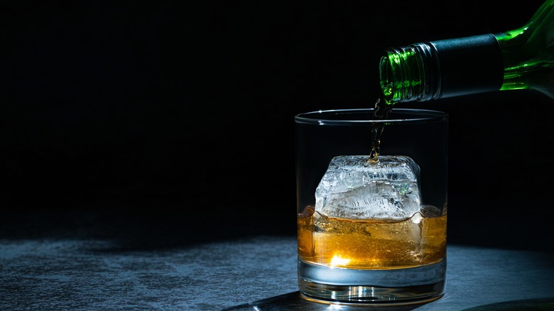 Liquid being poured from a green bottle into a glass with a large ice cube